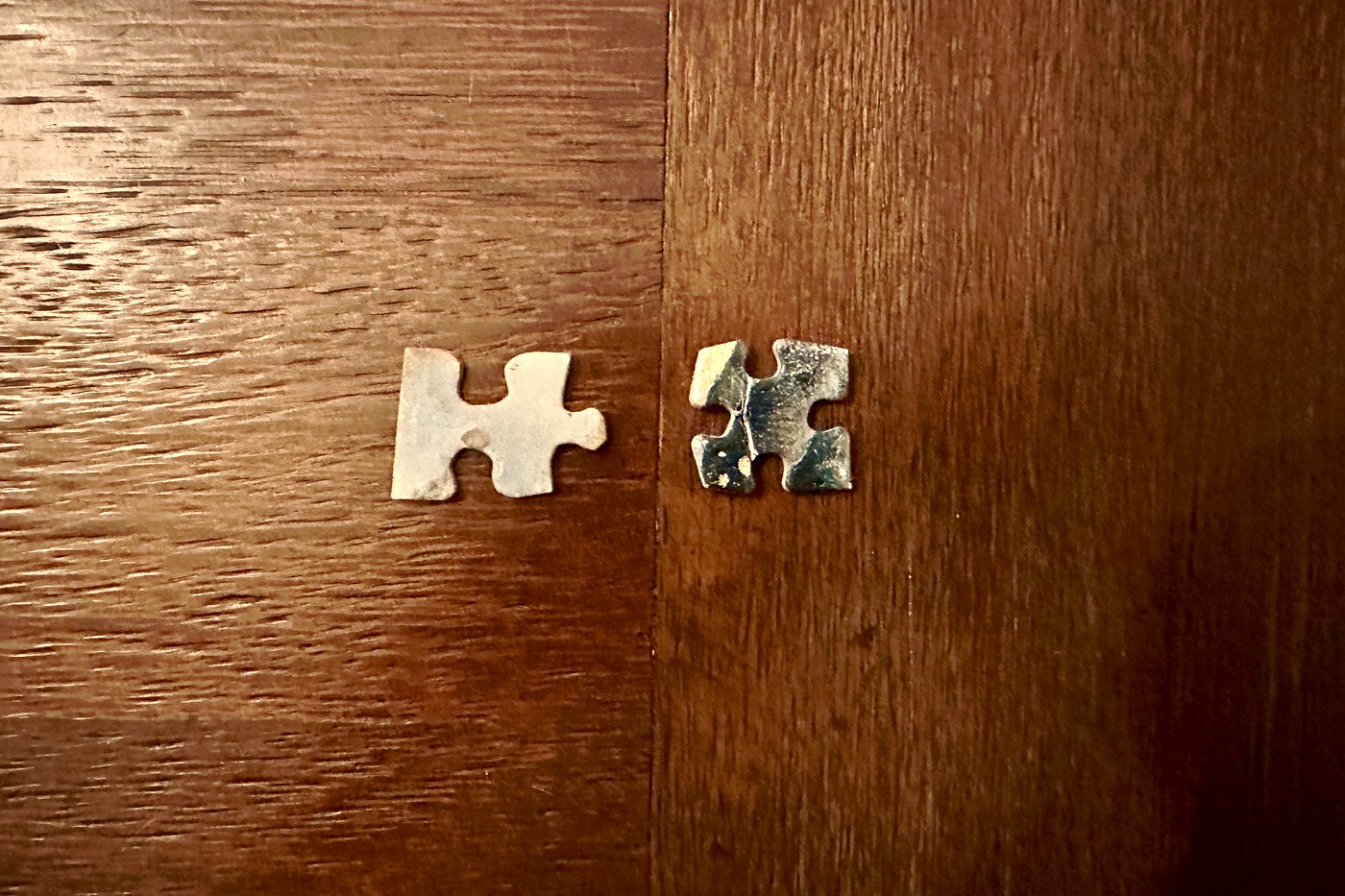 Two puzzle pieces on wood — one pale and worn, one darker, reaching toward each other across the grain